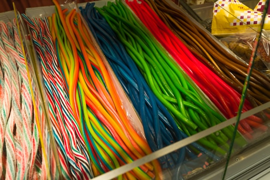 A variety of colorful candy sticks neatly arranged in a display. The candies are twisted into long strands with vibrant colors including pink, red, blue, green, yellow, and brown. Each group of candy has distinct color patterns and is separated in its own section within a glass display case.