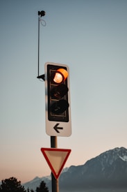 A traffic light with an illuminated amber light is mounted on a pole. Above it, a camera or sensor is attached to a tall antenna. Below the traffic light is a yield sign with a red border. A scenic backdrop of mountains is visible under a clear sky.