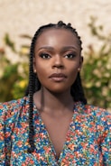 A woman with braided hair is standing in front of a background featuring greenery. She has a calm and composed expression on her face and is wearing a colorful floral-patterned top.