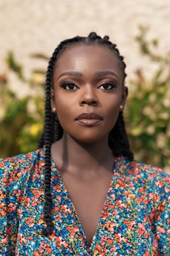 A woman with braided hair is standing in front of a background featuring greenery. She has a calm and composed expression on her face and is wearing a colorful floral-patterned top.