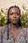 A woman with braided hair is standing in front of a background featuring greenery. She has a calm and composed expression on her face and is wearing a colorful floral-patterned top.