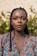 A woman with braided hair is standing in front of a background featuring greenery. She has a calm and composed expression on her face and is wearing a colorful floral-patterned top.