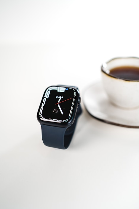A sleek smartwatch with a black band displays the time against a clean white background. Next to it is a white ceramic cup filled with coffee on a matching saucer.