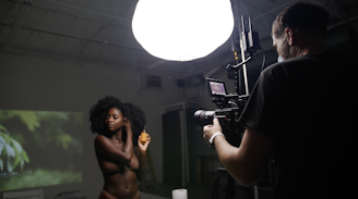 A dimly lit studio environment with a bright overhead light illuminating a photographer and a model. The model, a woman with natural hair, is posing and holding a spray bottle. The photographer is operating a professional camera mounted on a tripod, capturing the scene.