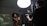 A dimly lit studio environment with a bright overhead light illuminating a photographer and a model. The model, a woman with natural hair, is posing and holding a spray bottle. The photographer is operating a professional camera mounted on a tripod, capturing the scene.