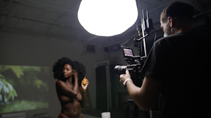 A dimly lit studio environment with a bright overhead light illuminating a photographer and a model. The model, a woman with natural hair, is posing and holding a spray bottle. The photographer is operating a professional camera mounted on a tripod, capturing the scene.