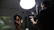 A dimly lit studio environment with a bright overhead light illuminating a photographer and a model. The model, a woman with natural hair, is posing and holding a spray bottle. The photographer is operating a professional camera mounted on a tripod, capturing the scene.