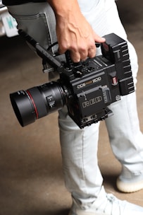 A person is holding a professional RED digital cinema camera with a large lens and multiple attachments. The camera has visible branding, buttons, and handles, indicating it is designed for high-quality film production. The person is wearing light-colored jeans and sneakers.