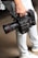 A person is holding a professional RED digital cinema camera with a large lens and multiple attachments. The camera has visible branding, buttons, and handles, indicating it is designed for high-quality film production. The person is wearing light-colored jeans and sneakers.