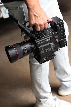 A person is holding a professional RED digital cinema camera with a large lens and multiple attachments. The camera has visible branding, buttons, and handles, indicating it is designed for high-quality film production. The person is wearing light-colored jeans and sneakers.
