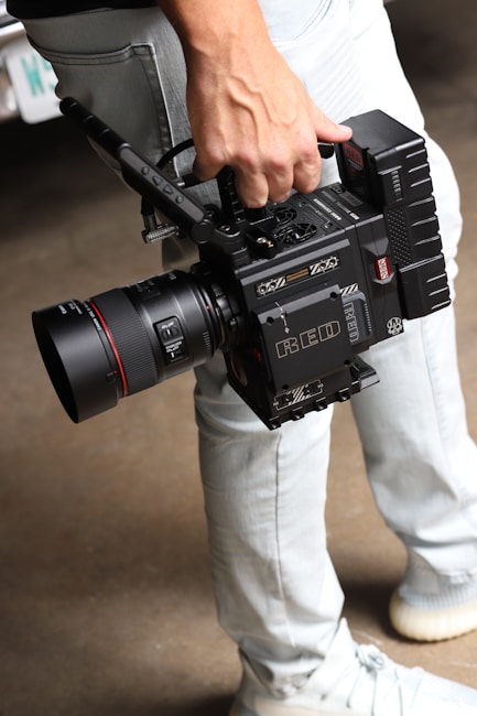A person is holding a professional RED digital cinema camera with a large lens and multiple attachments. The camera has visible branding, buttons, and handles, indicating it is designed for high-quality film production. The person is wearing light-colored jeans and sneakers.