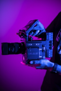 A person holds a professional digital camera with a large lens. The camera is branded as RED and features a sleek, modern design with various dials and grips. The lighting casts a magenta and blue hue over the scene, creating a dramatic effect.