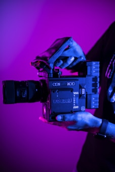 A person holds a professional digital camera with a large lens. The camera is branded as RED and features a sleek, modern design with various dials and grips. The lighting casts a magenta and blue hue over the scene, creating a dramatic effect.