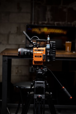 A high-quality video camera with an orange and black design is mounted on a professional tripod. The background includes a wooden table with a beer bottle and a pint glass. The setting appears to be a dimly lit room with a rustic or industrial aesthetic, highlighted by a blurred television screen displaying an abstract pattern.