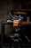 A high-quality video camera with an orange and black design is mounted on a professional tripod. The background includes a wooden table with a beer bottle and a pint glass. The setting appears to be a dimly lit room with a rustic or industrial aesthetic, highlighted by a blurred television screen displaying an abstract pattern.