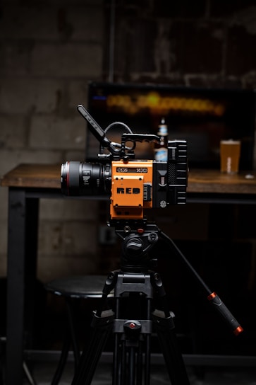 A high-quality video camera with an orange and black design is mounted on a professional tripod. The background includes a wooden table with a beer bottle and a pint glass. The setting appears to be a dimly lit room with a rustic or industrial aesthetic, highlighted by a blurred television screen displaying an abstract pattern.