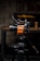 A high-quality video camera with an orange and black design is mounted on a professional tripod. The background includes a wooden table with a beer bottle and a pint glass. The setting appears to be a dimly lit room with a rustic or industrial aesthetic, highlighted by a blurred television screen displaying an abstract pattern.