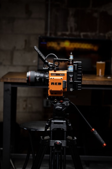 A high-quality video camera with an orange and black design is mounted on a professional tripod. The background includes a wooden table with a beer bottle and a pint glass. The setting appears to be a dimly lit room with a rustic or industrial aesthetic, highlighted by a blurred television screen displaying an abstract pattern.