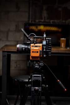 A high-quality video camera with an orange and black design is mounted on a professional tripod. The background includes a wooden table with a beer bottle and a pint glass. The setting appears to be a dimly lit room with a rustic or industrial aesthetic, highlighted by a blurred television screen displaying an abstract pattern.
