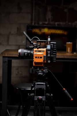 A high-quality video camera with an orange and black design is mounted on a professional tripod. The background includes a wooden table with a beer bottle and a pint glass. The setting appears to be a dimly lit room with a rustic or industrial aesthetic, highlighted by a blurred television screen displaying an abstract pattern.