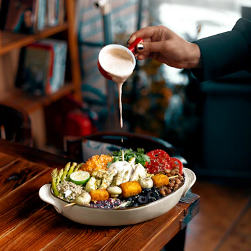 A hand is pouring a creamy white sauce over a bowl filled with a vibrant assortment of fresh vegetables and other components, including sliced cucumbers, tomatoes, mushrooms, beans, and possibly cheese. The setting appears to be a cozy, wooden table, and there's a blurred background of shelves and books.