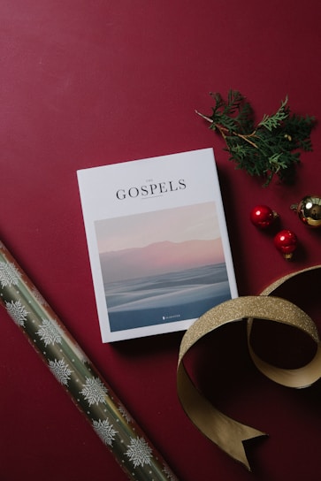 A book titled 'The Gospels' lies flat on a burgundy background. Nearby, there is a sprig of green foliage and three decorative red and gold ornaments, adding a festive touch. A length of gold ribbon is artfully placed beside them. To the left, a roll of wrapping paper with snowflake patterns is partially visible.