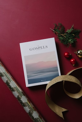 A book titled 'The Gospels' lies flat on a burgundy background. Nearby, there is a sprig of green foliage and three decorative red and gold ornaments, adding a festive touch. A length of gold ribbon is artfully placed beside them. To the left, a roll of wrapping paper with snowflake patterns is partially visible.