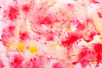 A vibrant abstract watercolor painting featuring a blend of pink, red, and yellow hues. The colors appear as swirling patterns and blotches, creating a dynamic and artistic effect. The texture is soft and fluid, with areas of light and shade adding depth.
