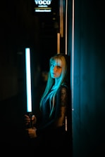 A person with long hair and tattoos is holding a blue light stick, standing against a wall with a calm, contemplative expression. The scene is dimly lit with an artificial blue glow, creating a mysterious atmosphere. The background is dark, with some signage partially visible.