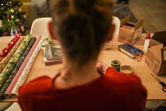 A person is sitting at a wooden table with various items related to gift wrapping. Rolls of wrapping paper with holiday-themed patterns are neatly arranged, alongside cardboard boxes, a notebook or journal, and some tape. In the background, a decorated Christmas tree is partially visible, indicating a festive atmosphere.