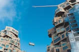 Two modern buildings with unique, abstract architectural designs featuring varying levels and protrusions. The structures are composed of light brown stone-like materials with numerous windows reflecting light. A construction crane is hoisting or lowering an object or platform between the buildings under a clear blue sky.