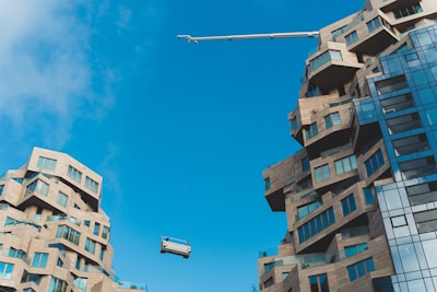 Two modern buildings with unique, abstract architectural designs featuring varying levels and protrusions. The structures are composed of light brown stone-like materials with numerous windows reflecting light. A construction crane is hoisting or lowering an object or platform between the buildings under a clear blue sky.