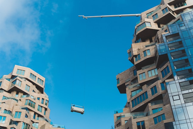 Two modern buildings with unique, abstract architectural designs featuring varying levels and protrusions. The structures are composed of light brown stone-like materials with numerous windows reflecting light. A construction crane is hoisting or lowering an object or platform between the buildings under a clear blue sky.