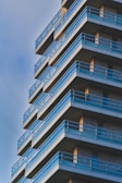 A modern high-rise building with a geometric design featuring multiple balconies with blue railings. The structure is composed of brick and metal elements, with each floor slightly protruding outwards. The sky is clear, adding a serene backdrop to the architectural design.