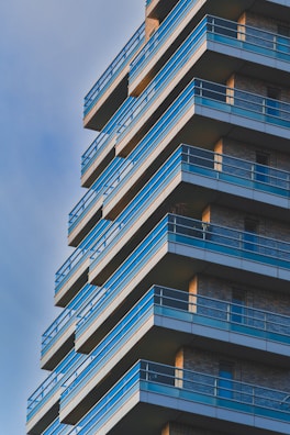 A modern high-rise building with a geometric design featuring multiple balconies with blue railings. The structure is composed of brick and metal elements, with each floor slightly protruding outwards. The sky is clear, adding a serene backdrop to the architectural design.