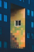 A building facade features a window set within a striking array of colorful tiles. The tiles form a rectangular section with a gradient pattern of greens, yellows, oranges, and browns, creating an eye-catching contrast against the surrounding dark brick wall.