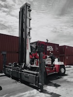 A large industrial vehicle designed for lifting and moving heavy objects is positioned next to several stacked shipping containers. A person wearing a protective suit and helmet is climbing onto the machine, which is painted primarily red and white. The overcast sky adds a muted backdrop to the scene.