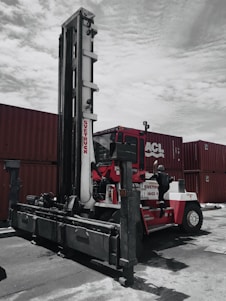 A large industrial vehicle designed for lifting and moving heavy objects is positioned next to several stacked shipping containers. A person wearing a protective suit and helmet is climbing onto the machine, which is painted primarily red and white. The overcast sky adds a muted backdrop to the scene.