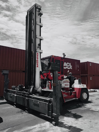 A large industrial vehicle designed for lifting and moving heavy objects is positioned next to several stacked shipping containers. A person wearing a protective suit and helmet is climbing onto the machine, which is painted primarily red and white. The overcast sky adds a muted backdrop to the scene.
