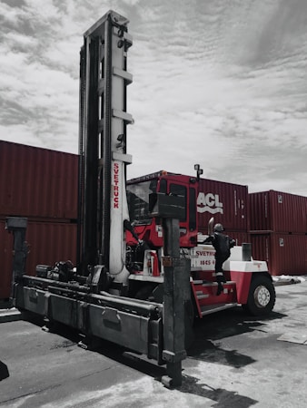A large industrial vehicle designed for lifting and moving heavy objects is positioned next to several stacked shipping containers. A person wearing a protective suit and helmet is climbing onto the machine, which is painted primarily red and white. The overcast sky adds a muted backdrop to the scene.