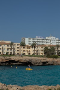 A coastline scene featuring a hotel named 'Globales' alongside other buildings with a sandy beige exterior. The foreground showcases a rugged shoreline with a small cave, and a vibrant blue sea. A person is seen in a bright yellow pedal boat on the water, adding a touch of activity to the tranquil setting.