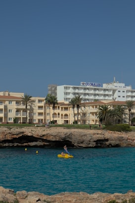 A coastline scene featuring a hotel named 'Globales' alongside other buildings with a sandy beige exterior. The foreground showcases a rugged shoreline with a small cave, and a vibrant blue sea. A person is seen in a bright yellow pedal boat on the water, adding a touch of activity to the tranquil setting.