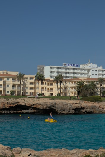 A coastline scene featuring a hotel named 'Globales' alongside other buildings with a sandy beige exterior. The foreground showcases a rugged shoreline with a small cave, and a vibrant blue sea. A person is seen in a bright yellow pedal boat on the water, adding a touch of activity to the tranquil setting.