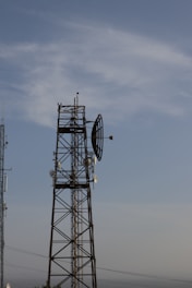 A tall metal communications tower with multiple antennas is set against a clear blue sky. The structure is composed of a lattice framework supporting various dishes and antennas.