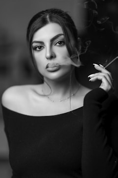 A woman with dark hair and dramatic makeup is holding a cigarette. Smoke curls around her face in an artistic manner. She wears an off-the-shoulder top, and the image has a moody, stylish aura.