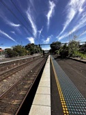 A railway platform extends into the distance beneath a clear sky with dramatic, wispy clouds. Parallel train tracks run beside the platform, and there are overhead electrical wires. Lush green trees and bushes line the area, while a blue and yellow tactile paving strip runs along the edge of the platform.