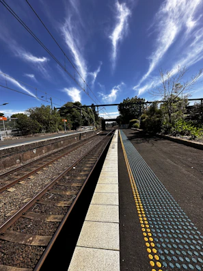 A railway platform extends into the distance beneath a clear sky with dramatic, wispy clouds. Parallel train tracks run beside the platform, and there are overhead electrical wires. Lush green trees and bushes line the area, while a blue and yellow tactile paving strip runs along the edge of the platform.
