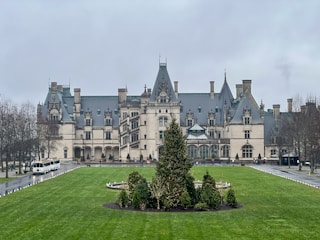 A large, elaborate mansion with a grand architectural design, featuring multiple chimneys and ornate detailing. The building is surrounded by well-manicured lawns and a circular driveway with transport vehicles parked. Overcast skies and the muted tones of winter vegetation contribute to the imposing presence of the structure.