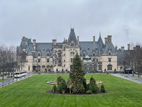 A large, elaborate mansion with a grand architectural design, featuring multiple chimneys and ornate detailing. The building is surrounded by well-manicured lawns and a circular driveway with transport vehicles parked. Overcast skies and the muted tones of winter vegetation contribute to the imposing presence of the structure.