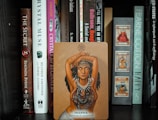 A shelf filled with various books, featuring a prominent card in the center depicting a woman with her arms crossed above her head, adorned with necklaces and symbols. The card reads 'Inanna - Believe in yourself.'